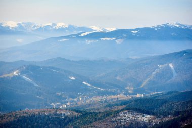 Katmanlı dağ sıralarının havadan görünüşü kısmen karla kaplı. Yoğun ormanlar ve dağınık yerleşimlerle dolu vadiler, uzak karlı zirveler soluk mavi gökyüzünün altında görkemli bir şekilde yükselirken.