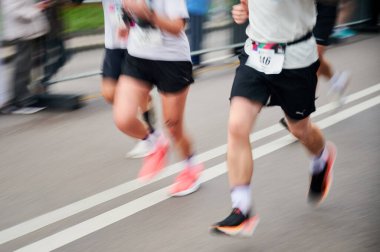 Hareket halindeki koşucular, rengarenk ayakkabılar ve atletik giysiler ile caddede koşuşturuyor. Kararlılıkla yarışırken dinamik ve enerjik bir sahne yaratıyorlar..