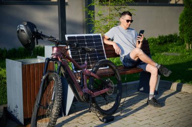 Bisikletçi, dışarıda elektrikli dağ bisikletini şarj etmek için güneş panelini kullanıyor. Güneş gözlüklü adam bankta oturur, telefon eder, tişört giyer, şort giyer, açık havada güneşlenmeyi sever..
