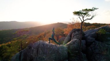 Yoga ve meditasyon yapan bir kadın akşamları dev bir kayanın tepesinde. Güzel günbatımı, sonbahar ormanları, arka planda kayalar ve tepeler. Hava aracı görünümü