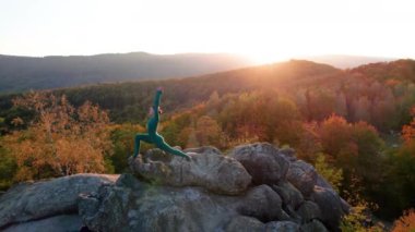 Yoga ve meditasyon yapan bir kadın akşamları dev bir kayanın tepesinde. Güzel günbatımı, sonbahar ormanları, arka planda kayalar ve tepeler. Hava aracı görünümü