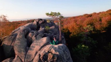 Yoga ve meditasyon yapan bir kadın akşamları dev bir kayanın tepesinde. Güzel günbatımı, sonbahar ormanları, arka planda kayalar ve tepeler. Hava aracı görünümü