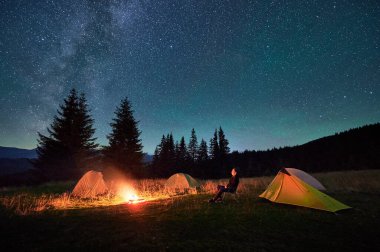 Person hiker having a rest by campfire, surrounded by glowing tents under magnificent starry sky. Milky Way stretches above, creating serene and magical atmosphere in peaceful mountain wilderness. clipart