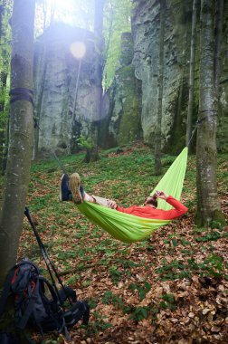Parlak yeşil hamakta dinlenen kişi kayaların yakınındaki yemyeşil ormandaki iki ağacın arasına bağlanmış. Turist adam içkisini yudumluyor, doğa ile çevrili huzurlu anın tadını çıkarıyor. Sırt çantası ve teçhizat yakında..