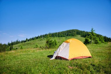 Bright yellow tent on grassy hillside, overlooking breathtaking panorama of blue-tinged mountains. Young pine trees under clear blue sky, creating serene and picturesque camping spot. clipart