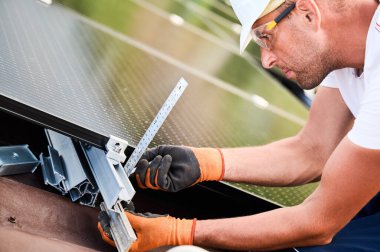 Fotovoltaik etkilerle elektrik üretmek için evin çatısına güneş paneli inşa eden işçiler. Kurulum için montaj ekipmanlarını ölçmek için cetvel kullanan adam.