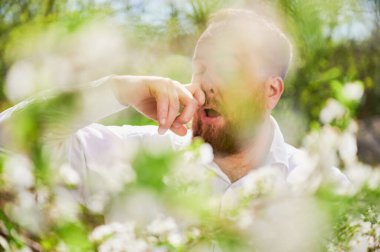 Baharda baharda çiçek açan bahçede mevsimsel alerji yüzünden acı çeken bir adam. Çiçek açan ağacın önünde hapşıran sakallı adamı yakından çek. Bahar alerjisi konsepti.