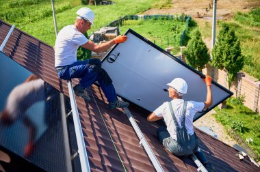 İşçiler evin çatısına güneş paneli inşa ediyorlar. Dışarıda fotovoltaik güneş modülü kuran kask takan iki adam. Alternatif, yeşil ve yenilenebilir enerji üretimi kavramı.
