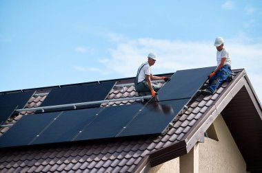 İşçiler evin çatısına güneş paneli inşa ediyorlar. Dışarıda fotovoltaik güneş modülü kuran kask takan iki adam. Alternatif, yeşil ve yenilenebilir enerji üretimi kavramı.