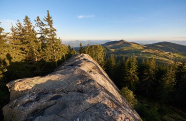 Stunning view from top of rocky outcrop overlooking Pysanyi Kamin in Carpathians, Ukraine. Background reveals rolling green hills, dense forests, and distant mountain ranges under clear blue sky. clipart