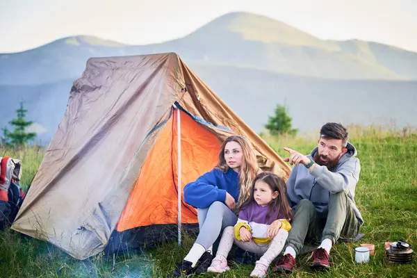 Üç kişilik aile çimenli dağ kamp alanındaki turist çadırının önünde oturur. Anne ve çocuk dikkatlice bakarken baba uzaktaki bir şeyi işaret ediyor. Açık gökyüzünün altındaki puslu dağlar.