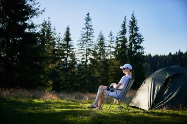 Kadın kampçı, fotoğraf makinesiyle çadırın yanında dinleniyor. Etrafı hep yeşil ağaçlarla çevrili ve yumuşak akşam ışığında yıkanan dişi turist, o anın huzurunu yakalıyor..