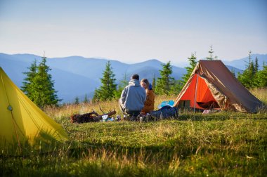 Sabahları iki çadırı olan manzaralı dağ kampı. Yuvarlanan dağların huzurlu manzarasının tadını çıkaran iki kampçının arka görüntüsü. Resimli gündoğumu manzarası, huzurlu ve rahat bir açık hava deneyimi yaratıyor..