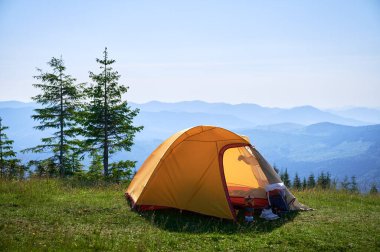 Bright yellow tent with camping gear on grassy hillside, overlooking breathtaking panorama of blue-tinged mountains. Young pine trees under clear blue sky, creating serene, picturesque camping spot. clipart