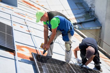 İşçiler evin metal çatısında fotovoltaik güneş paneli inşa ediyorlar. Erkek teknisyenler güneş modülünü açık havada elektrikli matkapla kuruyorlar. Alternatif enerji üretimi kavramı.