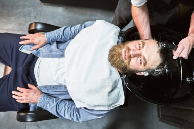 Kızıl saçlı adam saçını yıkatıyor. Erkek kuaför saçlarını derin temizlik ürünleriyle yıkıyor. Yakışıklı müşteri göğsünde havluyla rahatlıyor..