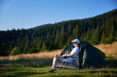 Kadın kampçı, fotoğraf makinesiyle çadırın yanında dinleniyor. Etrafı hep yeşil ağaçlarla çevrili ve yumuşak akşam ışığında yıkanan dişi turist, o anın huzurunu yakalıyor..
