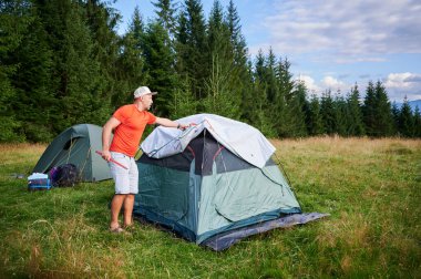 Yürüyüşçü otlak yamaçta turist çadırı kurdu. Çam ağaçları ve uzak dağlar parçalı bulutlu gökyüzünün altında.