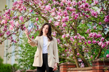 Bahar çiçekleri arasında akıllı telefonuyla selfie çeken bir kadın arka planda yumuşak bir odak ile çiçek açar..