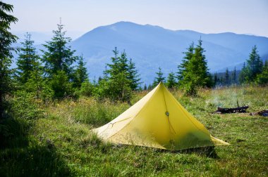 Gün batımının altında çimenli yamaçta turist çadırı. Etrafı genç çam ağaçlarıyla çevrili. Uzak dağların göz kamaştırıcı manzarası ve sabahları huzurlu ve pitoresk bir kamp yeri yaratıyor..