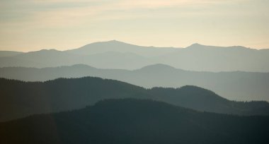 Yeşil ve gri yumuşak gölgelerdeki katmanlı dağların büyüleyici görüntüsü ince, sisli bir gökyüzünün altında derinlik ve sükunet hissi yaratıyor..