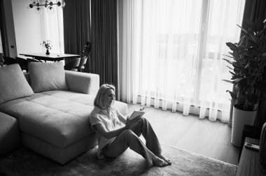 Kanepenin yanında oturan ve kitap okuyan genç bir kadın. Güzel bir kadın, ahşap zeminde dinleniyor. Kitapla oturan kadın pencerenin karşısında, doğal ışıkta. Siyah ve beyaz resim.