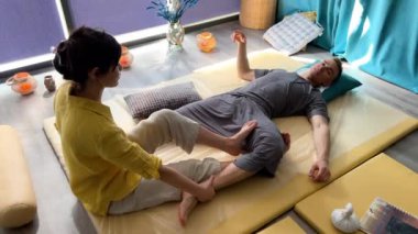 Masör Tayland yogası masajı yapıyor. Geleneksel masaj tedavisi uygulayan kadın terapist, erkek hastaların vücudunu esnetiyor. Müşteri paspasın üzerinde uzanıyor, sakin atmosferde el masajının keyfini çıkarıyor..