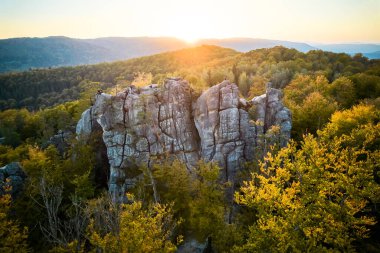 Güneşin hava manzarası engebeli kaya oluşumunun arkasında batar ve manzaranın üzerine sıcak bir ışıltı yayar. Sonbahar renklerinde ağaçlar kayaları çevreleyerek canlı bir kontrastlık yaratır. Dovbush Kayaları, Karpat Dağları, Ukrayna.
