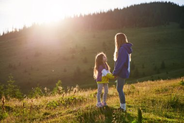 Genç bayan turist, gün batımında büyüleyici dağ manzarasına karşı gün ışığında kızıyla birlikte çayırda vakit geçiriyor. Neşeli an kaygısız eğlence ve doğal güzelliğin özünü yakalar..