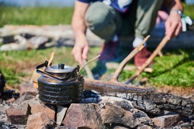 Etrafı taşlarla çevrili, kararmış çaydanlık ısıtıcısının yakın çekimi. Arka planda, sopayla ateş etmeye meyilli biri var. Çimenli çayır, kırsal kamp deneyimini ön plana çıkarıyor..
