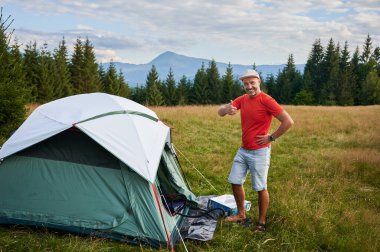 Erkek turist çimenli çayırlarda çadır kurup, yeşil çadırın içindeki şişme yatağı şişirmek için ayak pompasını kullanıyor. Arkaplanda çam ağaçları ve dağ parçalı bulutlu gökyüzünün altında.