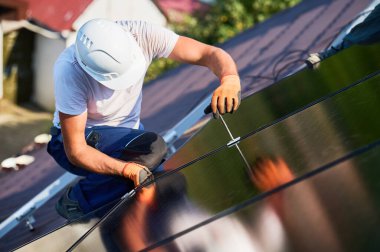 Evin çatısında fotovoltaik güneş paneli inşa eden bir işçi. Miğferlerde mühendis ve eldivenler dışarıda altıgen anahtarla güneş modülü kuruyor. Yenilenebilir enerji üretimi kavramı.