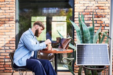 Adam dışarıdaki yuvarlak masada bir bardak buzlu kahveyle dizüstü bilgisayarda çalışıyor. Yakınlardaki güneş paneli, teknoloji ve sürdürülebilirliği birleştiren sürdürülebilir, modern çevre dostu çalışma alanını vurguluyor.