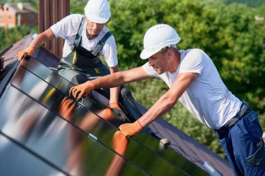 İşçiler evin çatısına fotovoltaik güneş paneli inşa ediyorlar. Miğferli teknisyenler ve eldivenli adamlar dışarıda büyücülerin yardımıyla güneş modülü kuruyorlar. Yenilenebilir enerji.