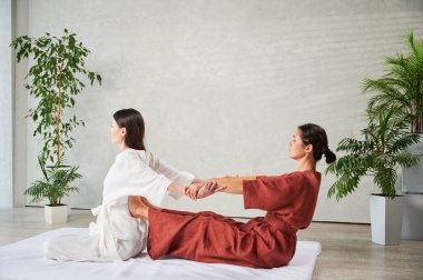 Masör Tayland yogası masajı yapıyor. Geleneksel masaj tedavisi uygulayan kadın terapist, kadın hastaların vücudunu geriyor. Müşteri paspasın üzerinde oturuyor, sakin bir atmosferde el masajının keyfini çıkarıyor..