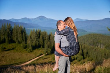 Çift, manzaralı dağ manzarasında samimi bir anı paylaşıyor. Erkek, kadını kucaklarken ve birbirlerinin gözlerine bakarken kendine yakın tutar. Bereketli yeşil orman ve açık mavi gökyüzünün altındaki uzak dağlar..