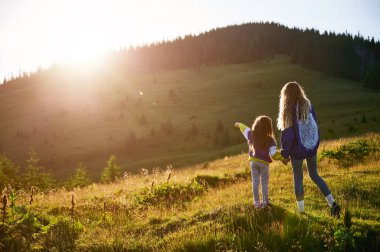 Genç bayan turist, gün batımında büyüleyici dağ manzarasına karşı gün ışığında kızıyla birlikte çayırda vakit geçiriyor. Neşeli an kaygısız eğlence ve doğal güzelliğin özünü yakalar..