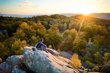 Ukrayna 'daki Karpat Dağları' ndaki Dovbush Kayalıkları 'ndaki kaya oluşumunun üzerinde oturan dağcı görüntüsü. Güneş batıyor, sonbahar ormanları ve uzak tepeler üzerinde parıldıyor, nefes kesici bir manzara yaratıyor..