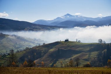 Sonbahar yapraklarıyla kaplı yuvarlanan tepelerin kısmen alçak sis örtüsüyle örtülü olduğu çarpıcı bir manzara. Karla kaplı dağlar arkaplanda, berrak mavi gökyüzünün altında görkemli bir şekilde yükselir.