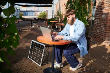 Adam dizüstü bilgisayarla çalışıyor ve dışarıdaki masada bir bardak buzlu kahveyle akıllı telefon kullanıyor. Yakınlardaki güneş paneli, teknoloji ve sürdürülebilirliği birleştiren sürdürülebilir, modern çevre dostu çalışma alanını vurguluyor.