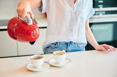 Lviv, Ukrayna - 26 Mayıs 2023: Bardağa su döken kadın resmi. Genç bayan iki kişilik çay yapıyor. İki fincanın üzerinde kırmızı çaydanlık tutan kadın..