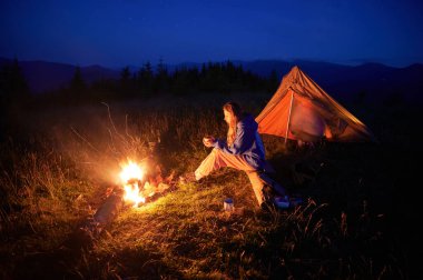 Kadın yürüyüşçü, sakin dağ manzarasıyla çevrili alacakaranlığın yumuşak parıltısıyla aydınlık çadırının yanında kamp ateşinin yanında oturur. Yukarıda açık bir gece gökyüzü sessiz vahşi doğaya huzur dolu bir ortam katıyor..