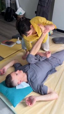 Masör Tayland yogası masajı yapıyor. Geleneksel masaj tedavisi uygulayan kadın terapist, erkek hastaların vücudunu esnetiyor. Müşteri paspasın üzerinde uzanıyor, sakin atmosferde el masajının keyfini çıkarıyor..