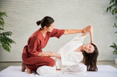 Masör Tayland yogası masajı yapıyor. Geleneksel masaj tedavisi uygulayan kadın terapist, kadın hastaların vücudunu geriyor. Müşteri paspasın üzerinde oturuyor, sakin bir atmosferde el masajının keyfini çıkarıyor..