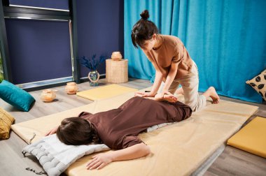 Masör Tayland yogası masajı yapıyor. Geleneksel masaj tedavisi uygulayan kadın terapist, kadın hastaların vücudunu geriyor. Müşteri paspasın üzerinde uzanıyor, sakin atmosferde el masajının keyfini çıkarıyor..