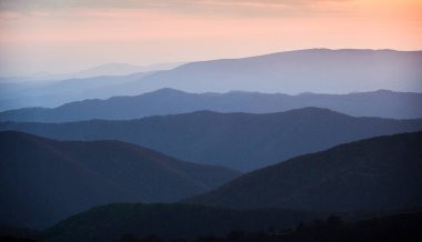 Alacakaranlıkta sakin bir sahne, yuvarlanan tepelerin gösterişli katmanları yumuşak mavi renklerle yıkanmış, gün batımının azalan ışığı uzak ufukta hafif turuncu ve pembe bir parıltı yayıyor..