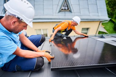 İşçiler evin çatısına güneş paneli inşa ediyorlar. Dışarıda fotovoltaik güneş modülü kuran kask takan iki adam. Alternatif, yeşil ve yenilenebilir enerji üretimi kavramı.
