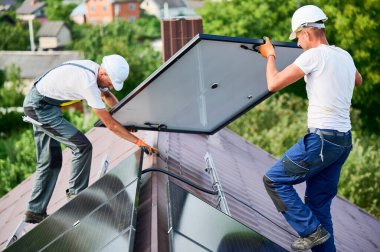 İşçiler evin çatısına güneş paneli inşa ediyorlar. Dışarıda fotovoltaik güneş modülü taşıyan kasklı iki adam var. Alternatif, yeşil ve yenilenebilir enerji üretimi kavramı.