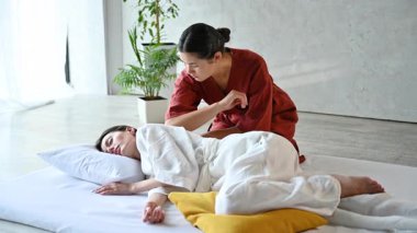 Masseuse making thai yoga massage. Woman therapist doing traditional massage treatment for female patient using elbow. Client lying on mat, meditating, enjoying hand massage in calm atmosphere.
