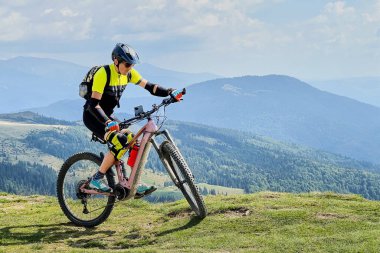 Dışarıda elektrikli dağ bisikleti süren bisikletçi. Erkek turist dağlarda otlak patika boyunca bisiklet sürüyor, başlık ve sırt çantası takıyor. Spor anlayışı, aktif eğlence ve doğa.